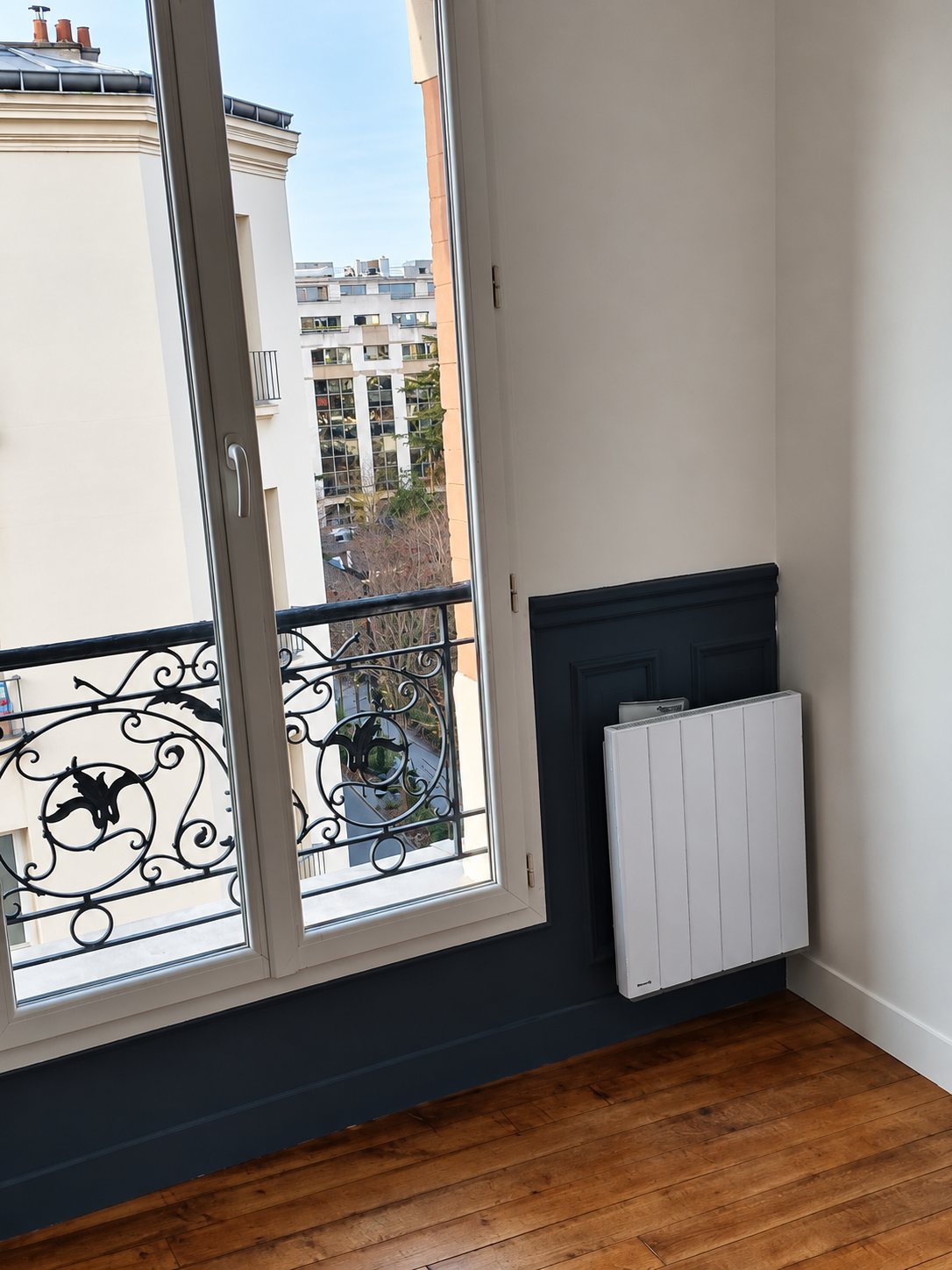 Fenêtre avec garde-corps en fer forgé, lambris bleu et radiateur blanc dans un appartement parisien rénové
